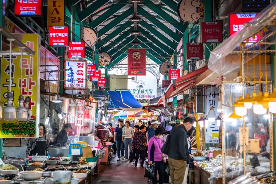 江陵中央市場有綠色上蓋的主通道、兩旁熟食與海產檔口及逛市場的人群